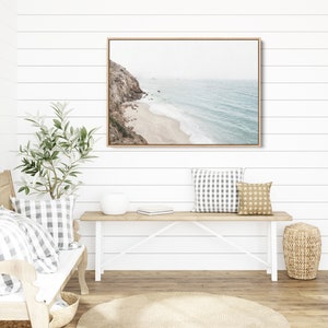 May include: A framed photograph of a coastal scene with a rocky cliff, sandy beach, and blue ocean waves. The photograph is hanging on a white shiplap wall above a white bench with two pillows.