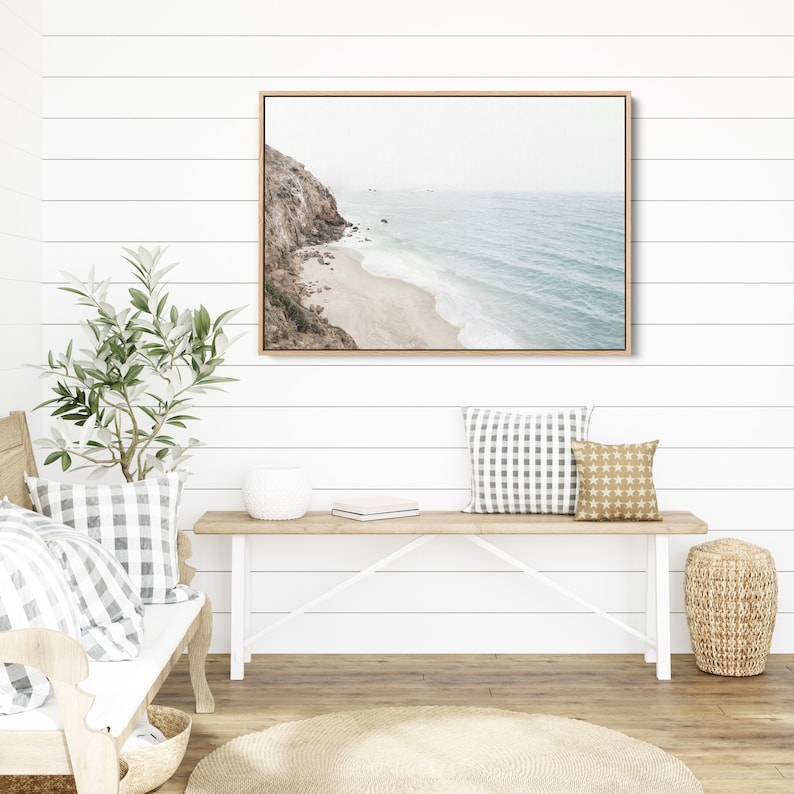 May include: A framed photograph of a coastal scene with a rocky cliff, sandy beach, and blue ocean waves. The photograph is hanging on a white shiplap wall above a white bench with two pillows.