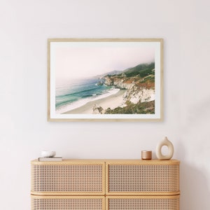 Peut inclure: Tirage encadr&eacute; d'un paysage c&ocirc;tier avec une plage, une eau turquoise, des falaises blanches et des collines verdoyantes sous un ciel brumeux. Le cadre est en bois clair. Il est expos&eacute; au-dessus d'un meuble en bois clair avec des panneaux tress&eacute;s.