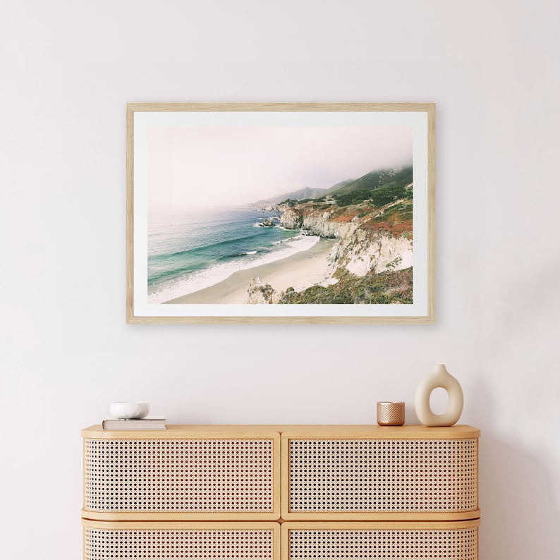 Peut inclure: Tirage encadr&eacute; d'un paysage c&ocirc;tier avec une plage, une eau turquoise, des falaises blanches et des collines verdoyantes sous un ciel brumeux. Le cadre est en bois clair. Il est expos&eacute; au-dessus d'un meuble en bois clair avec des panneaux tress&eacute;s.