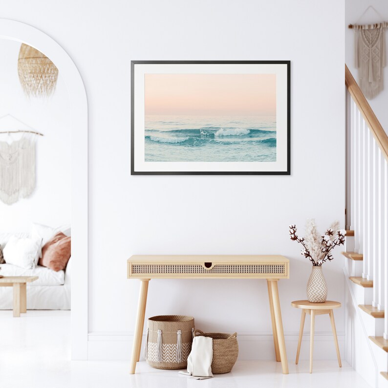 May include: A framed photograph of a seascape with waves crashing on the shore. The sky is a soft pink and the water is a light blue. The photo is hanging on a white wall above a wooden table with a woven basket.