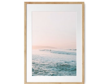 May include: A framed photograph of a surfer riding a wave in the ocean. The water is a light blue color and the sky is a soft pink. The photo is framed in a natural wood frame.