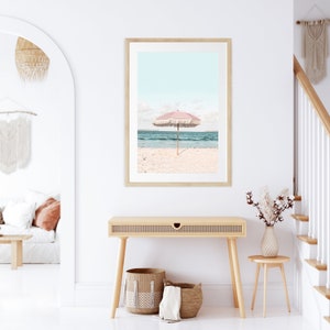 May include: A framed print of a pink beach umbrella on a white sandy beach with blue ocean water in the background.