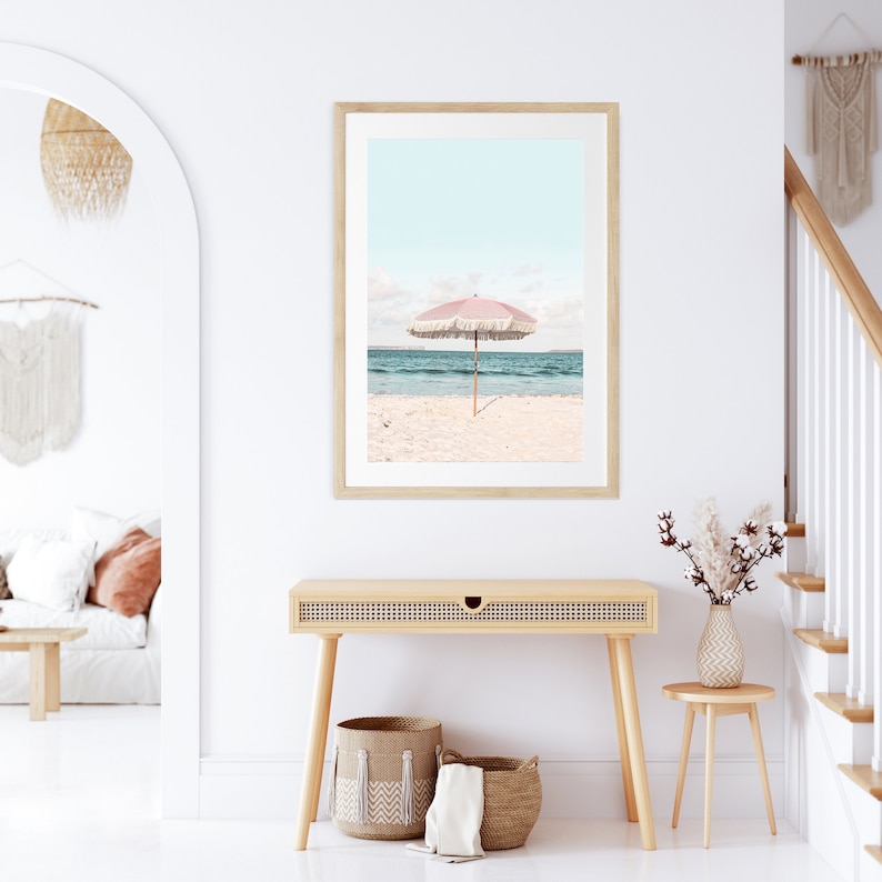 May include: A framed print of a pink beach umbrella on a white sandy beach with blue ocean water in the background.