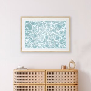 May include: A framed print of a blue and white abstract pattern of water ripples. The print is hanging on a white wall above a light brown dresser with two drawers.