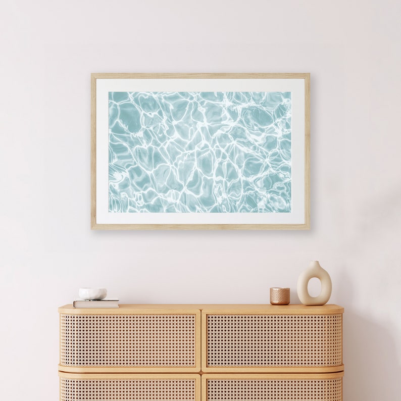 May include: A framed print of a blue and white abstract pattern of water ripples. The print is hanging on a white wall above a light brown dresser with two drawers.