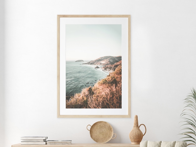 May include: A framed photograph of a coastal landscape with a view of a rocky coastline and the ocean. The photo is in a light wood frame and has a muted color palette.