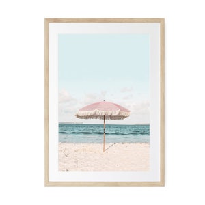 May include: A pink and white beach umbrella with fringe on a sandy beach with blue water in the background.