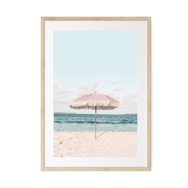 May include: A pink and white beach umbrella with fringe on a sandy beach with blue water in the background.
