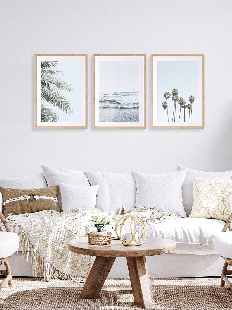 May include: Three framed prints of beach scenes. The left print shows a close-up of palm tree leaves. The center print shows a blue ocean with waves crashing on the shore. The right print shows a row of palm trees against a blue sky.
