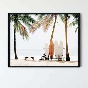 May include: Four surfboards leaning against a wooden rack on a sandy beach with palm trees in the background. The surfboards are white with a red and orange surfboard in the middle.