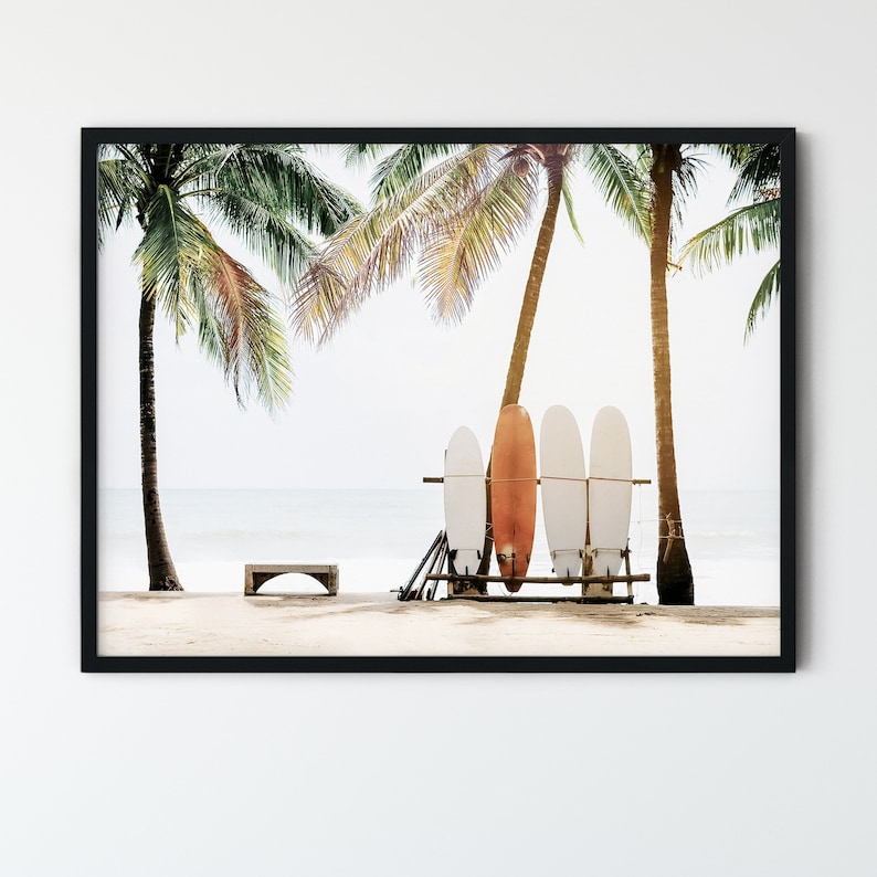 May include: Four surfboards leaning against a wooden rack on a sandy beach with palm trees in the background. The surfboards are white with a red and orange surfboard in the middle.