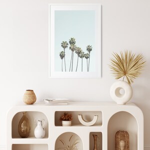 May include: White shelf unit with arched openings, featuring various decorative items, including vases, bowls, and dried plants. A framed print of palm trees against a light blue sky hangs above the shelf.