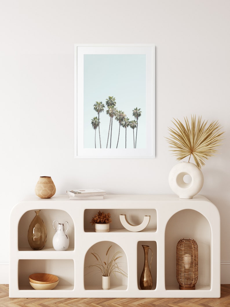 May include: White shelf unit with arched openings, featuring various decorative items, including vases, bowls, and dried plants. A framed print of palm trees against a light blue sky hangs above the shelf.