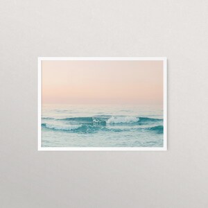 May include: A photograph of a seascape with soft, pink and blue hues. The ocean waves are gently rolling in the foreground, with a light pink sky in the background.