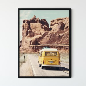 May include: A yellow vintage van drives down a paved road in a desert landscape. The van has a white roof and a surfboard strapped to the top. The van is driving past a large red rock formation.