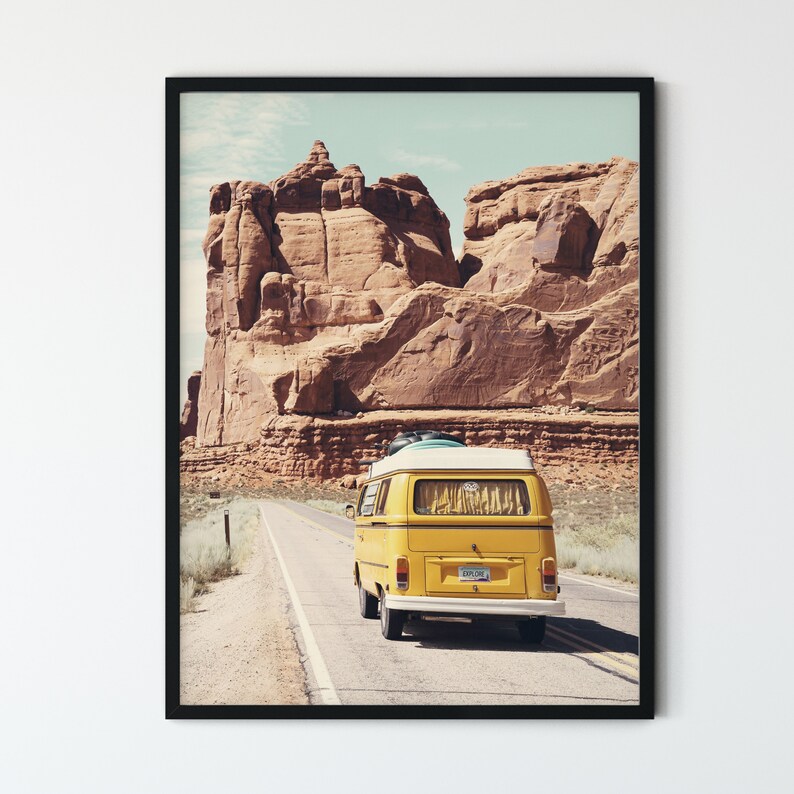 May include: A yellow vintage van drives down a paved road in a desert landscape. The van has a white roof and a surfboard strapped to the top. The van is driving past a large red rock formation.