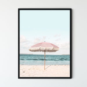 May include: A pink and white striped beach umbrella with white fringe on a white sandy beach with blue ocean water in the background.