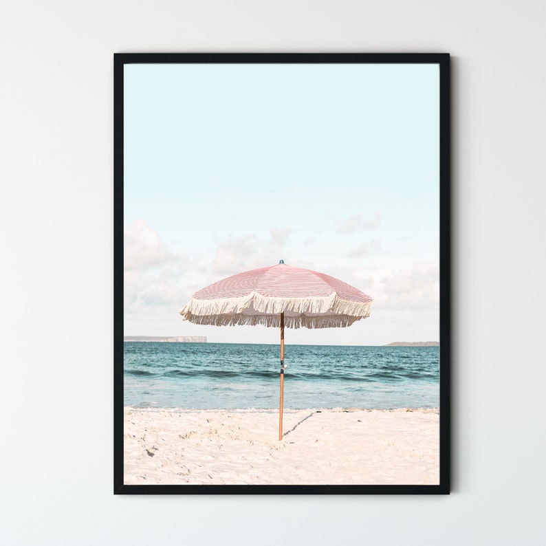 May include: A pink and white striped beach umbrella with white fringe on a white sandy beach with blue ocean water in the background.