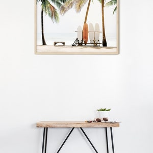 May include: A framed print of a beach scene with palm trees and surfboards. The surfboards are leaning against a wooden rack. The beach is sandy and the water is blue.