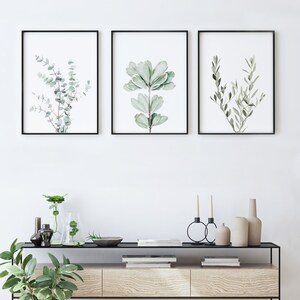 May include: Three framed botanical prints with black frames, featuring watercolor illustrations of different types of leaves. The prints are hanging on a white wall above a wooden console table with black metal legs. The table is decorated with vases, candles, and books.