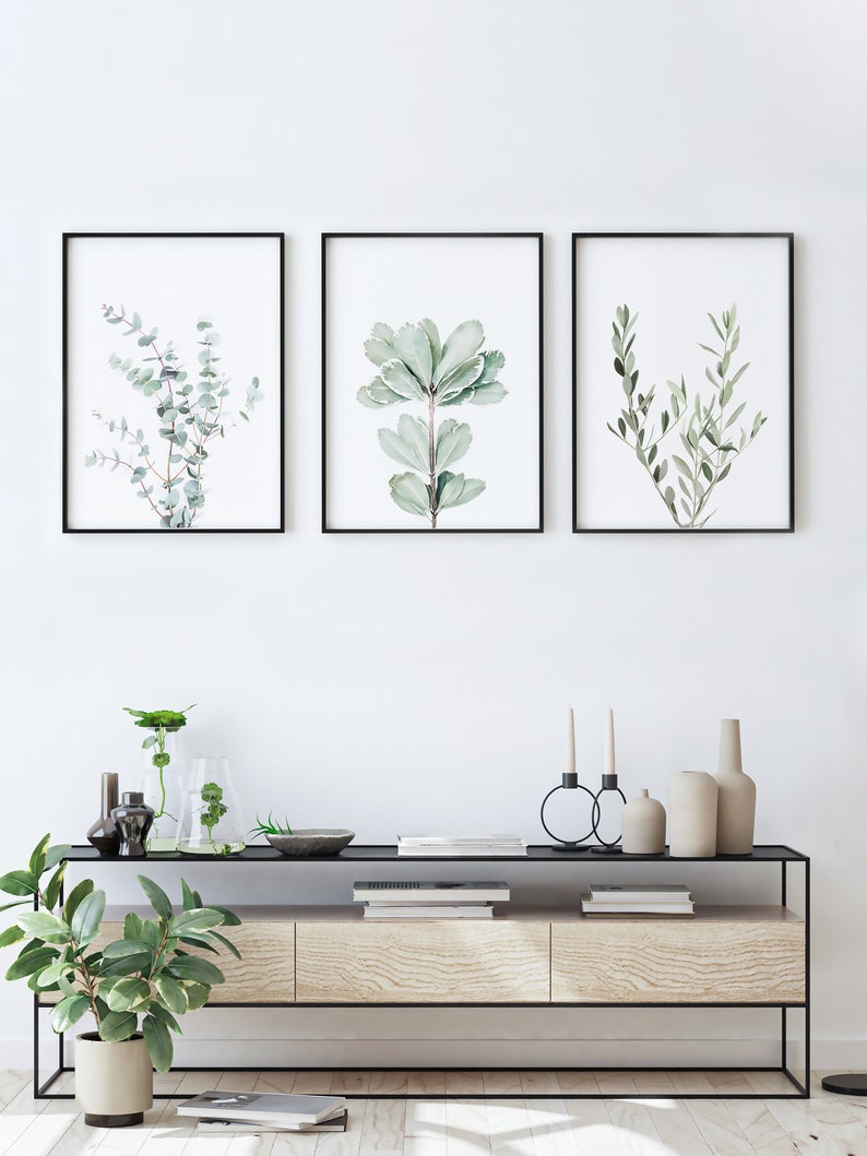 May include: Three framed botanical prints with black frames, featuring watercolor illustrations of different types of leaves. The prints are hanging on a white wall above a wooden console table with black metal legs. The table is decorated with vases, candles, and books.