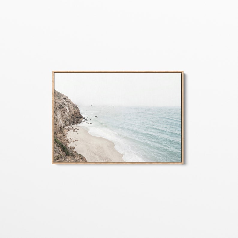 May include: A photograph of a rocky cliff overlooking a sandy beach and the ocean. The water is a light blue color and the sky is a hazy white.