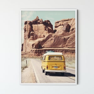 May include: A yellow vintage van drives down a paved road in a desert landscape with red rock formations in the background.