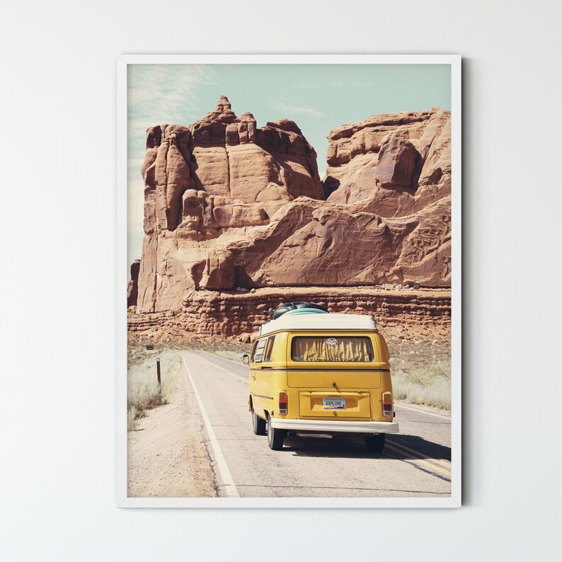 May include: A yellow vintage van drives down a paved road in a desert landscape with red rock formations in the background.