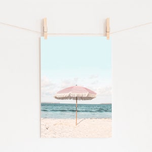 May include: A pink and white beach umbrella with fringe on a sandy beach with blue ocean water in the background.