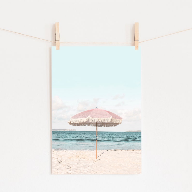 May include: A pink and white beach umbrella with fringe on a sandy beach with blue ocean water in the background.