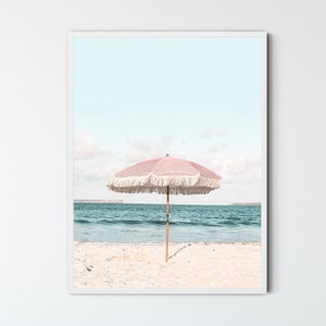 May include: A pink and white striped beach umbrella with white fringe stands on a white sandy beach with blue ocean water in the background.