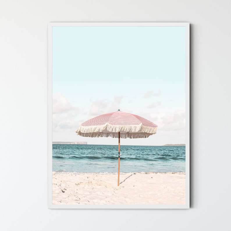 May include: A pink and white striped beach umbrella with white fringe stands on a white sandy beach with blue ocean water in the background.