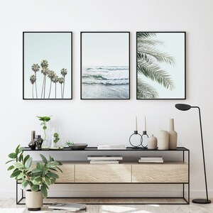 May include: Three framed prints with a beach theme. The left print shows palm trees against a light blue sky. The center print shows ocean waves crashing on the shore. The right print shows a close-up of palm fronds against a light blue sky.
