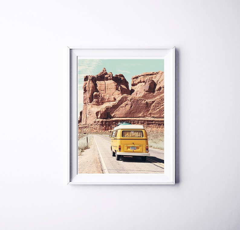May include: A yellow vintage van drives down a road in a desert landscape with red rock formations in the background.