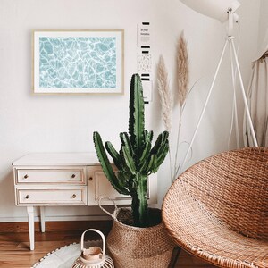 May include: A light blue framed print of rippling water, a potted cactus plant in a woven basket, a white floor lamp with a white shade, a white wicker chair, and a white dresser with three drawers.