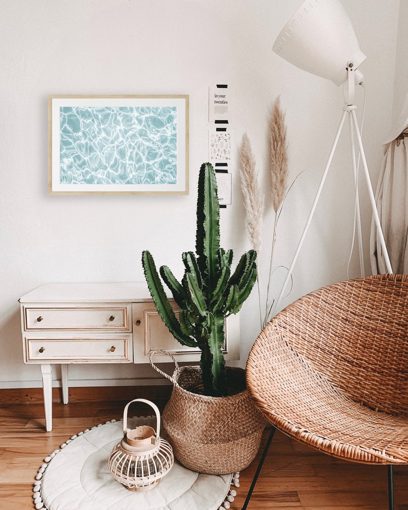 May include: A light blue framed print of rippling water, a potted cactus plant in a woven basket, a white floor lamp with a white shade, a white wicker chair, and a white dresser with three drawers.