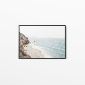 May include: A photograph of a rocky cliff overlooking a sandy beach and the ocean. The water is a light blue color and the waves are crashing on the shore. The sky is a light gray color and there are a few clouds in the distance.
