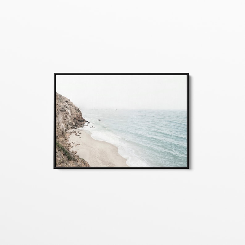 May include: A photograph of a rocky cliff overlooking a sandy beach and the ocean. The water is a light blue color and the waves are crashing on the shore. The sky is a light gray color and there are a few clouds in the distance.