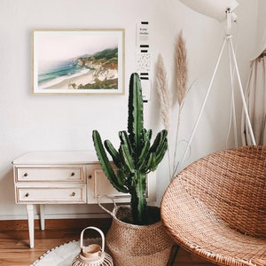 Peut inclure: Un cadre avec une impression de paysage c&ocirc;tier est accroch&eacute; &agrave; un mur blanc. Un grand cactus vert est dans un panier en osier. Une chaise en osier et une lampe blanche sont &eacute;galement dans la pi&egrave;ce. Un petit meuble blanc avec des tiroirs et un tapis rond compl&egrave;tent le d&eacute;cor.