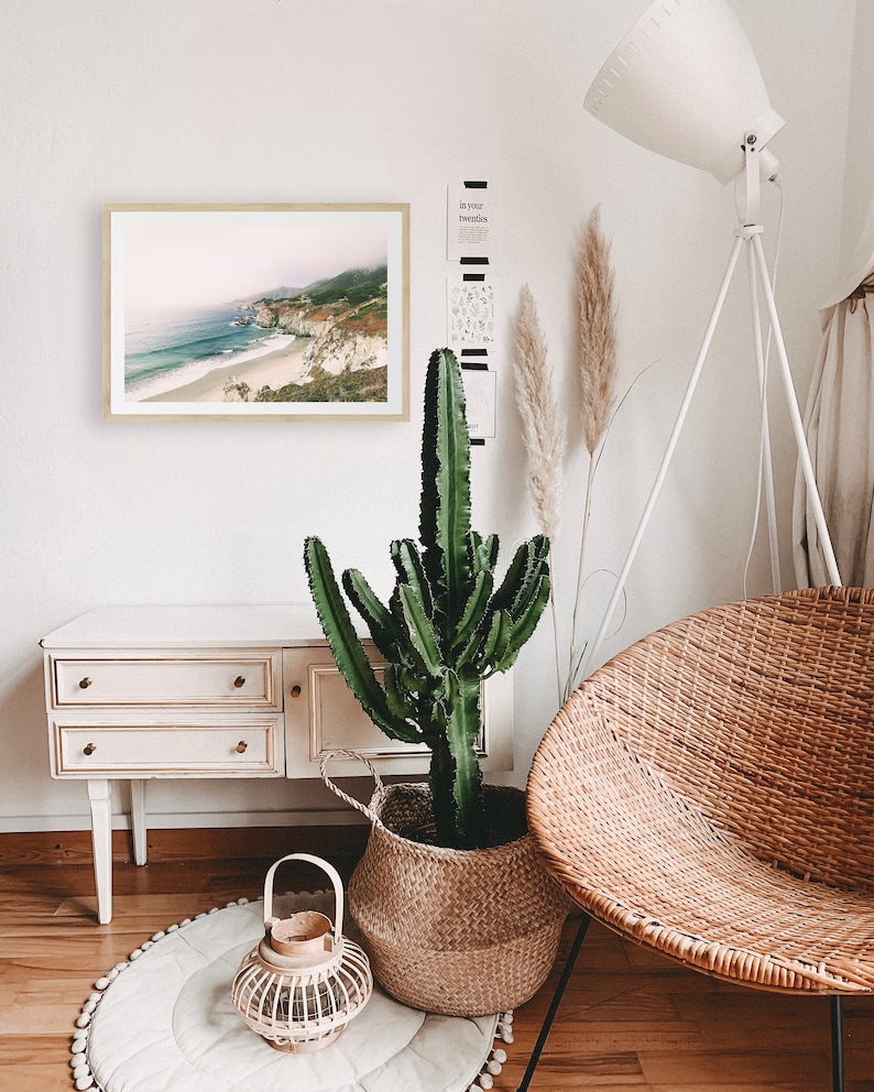 Peut inclure: Un cadre avec une impression de paysage c&ocirc;tier est accroch&eacute; &agrave; un mur blanc. Un grand cactus vert est dans un panier en osier. Une chaise en osier et une lampe blanche sont &eacute;galement dans la pi&egrave;ce. Un petit meuble blanc avec des tiroirs et un tapis rond compl&egrave;tent le d&eacute;cor.