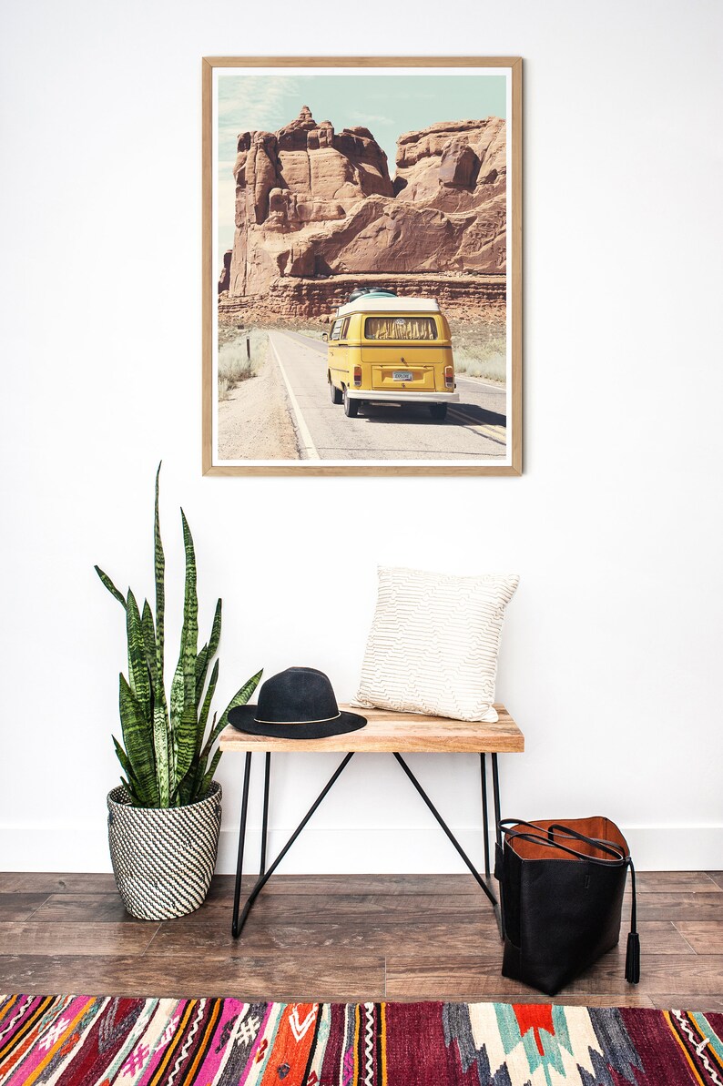 May include: A framed print of a yellow vintage van driving on a road through a desert landscape with red rock formations in the background.