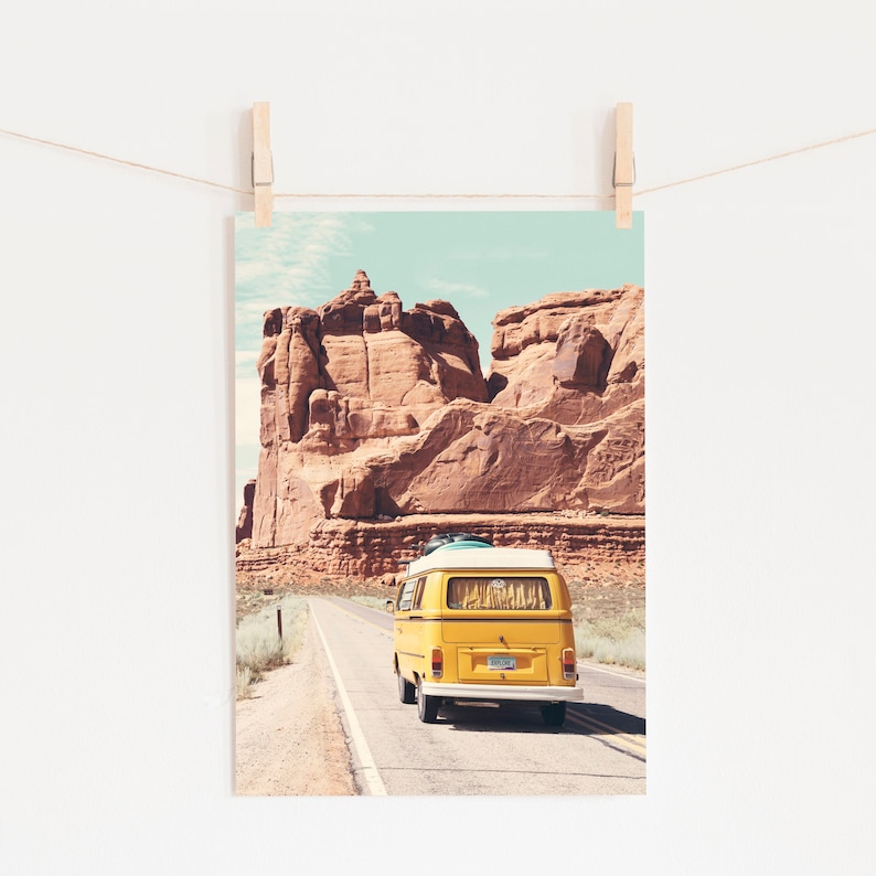 May include: A yellow vintage van drives down a road in a desert landscape with red rock formations in the background.