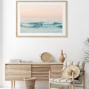 May include: A framed photograph of a seascape with waves crashing on the shore. The sky is a soft pink and the water is a light blue. The photo is hanging on a white wall above a wooden dresser.