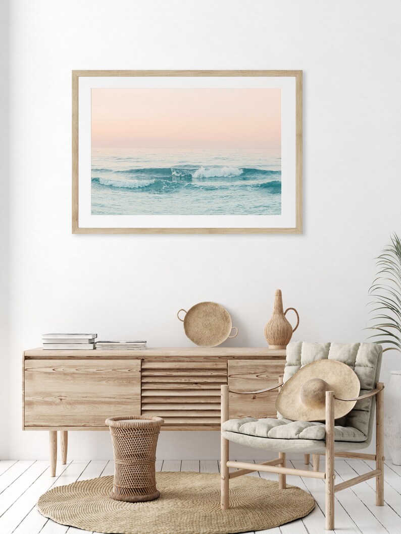 May include: A framed photograph of a seascape with waves crashing on the shore. The sky is a soft pink and the water is a light blue. The photo is hanging on a white wall above a wooden dresser.