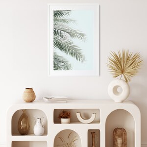 May include: White shelf unit with arched openings, featuring various decorative items, including a framed print of palm leaves, a vase with dried palm fronds, a wooden bowl, and a ceramic vase.