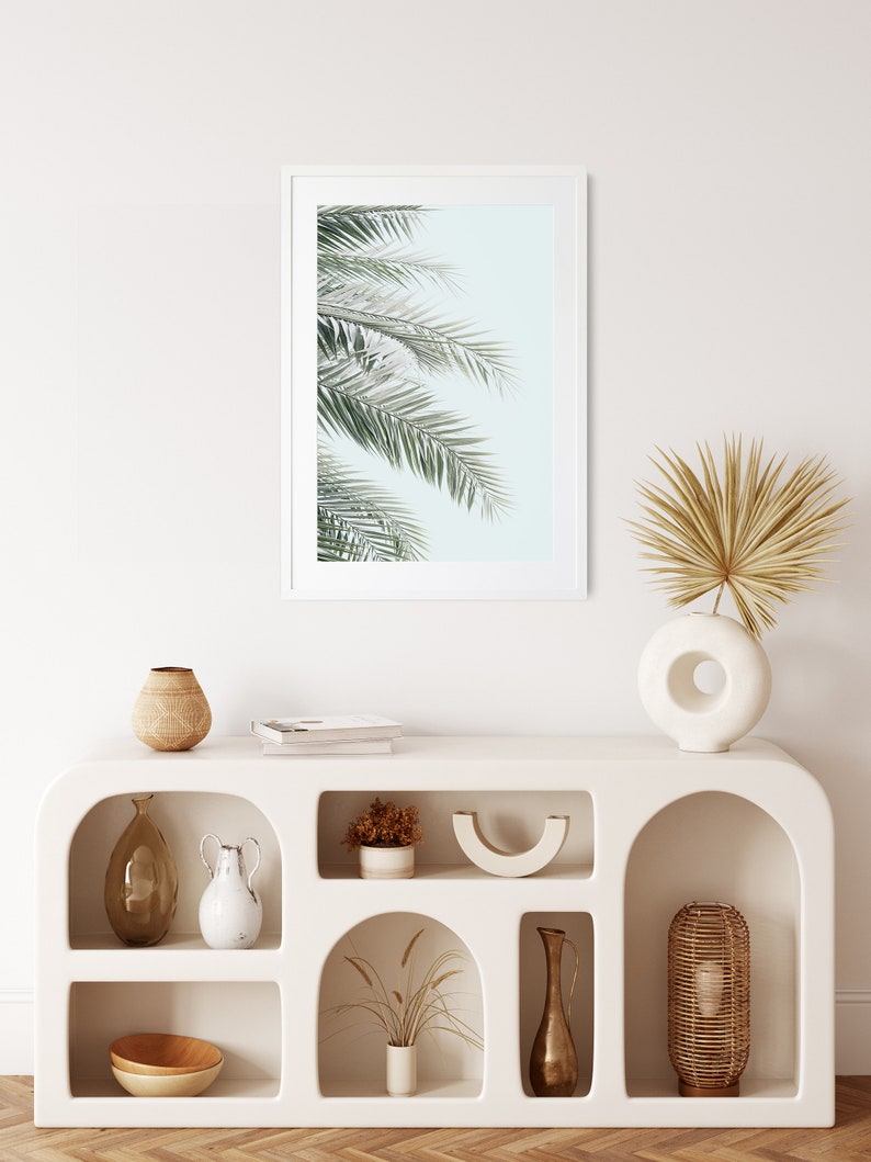 May include: White shelf unit with arched openings, featuring various decorative items, including a framed print of palm leaves, a vase with dried palm fronds, a wooden bowl, and a ceramic vase.