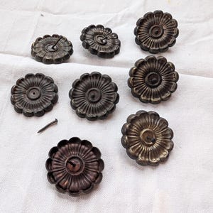May include: A collection of eight antique bronze-colored flower-shaped drawer pulls. Each pull has a central hole and a detailed petal design. A small nail is also present. The pulls are arranged on a white fabric surface.