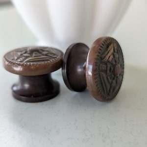 May include: Two dark brown wooden cabinet knobs with a circular face featuring a carved geometric star pattern. The knobs have a smooth, polished finish and a cylindrical base, showcasing a detailed relief design on the front surface of each piece.