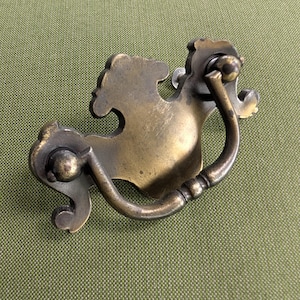 May include: A brass-toned metal drawer pull featuring a decorative backplate with curved, ornate edges and a swinging, U-shaped handle attached to two circular mounting points. The metal surface displays a weathered, aged patina finish.